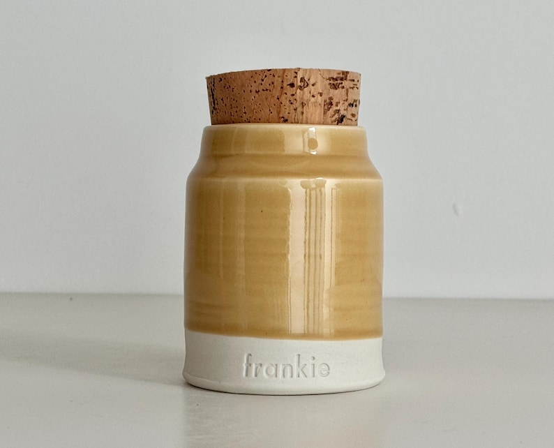 Image shows a collared shape urn with cork lid. It has an orange / wheat colored glazed upper section, and white unglazed clay lower portion. There is a custom name stamped into the clay