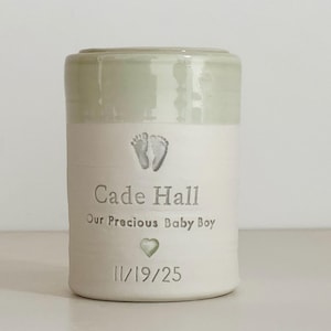 A sage green glaze and white porcelain keepsake infant urn. It features a baby footprint, the custom name, a phrase, a heart, and the date.
