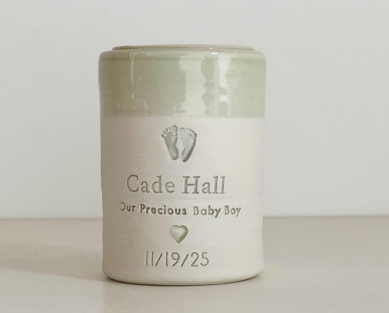 A sage green glaze and white porcelain keepsake infant urn. It features a baby footprint, the custom name, a phrase, a heart, and the date.
