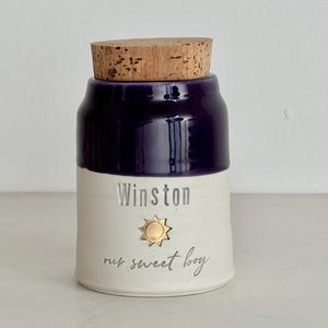 Image shows a collared shape urn with cork lid. It has a dark purple glazed upper section, and white unglazed clay lower portion. There is a gold sun stamp with name  and custom phrase