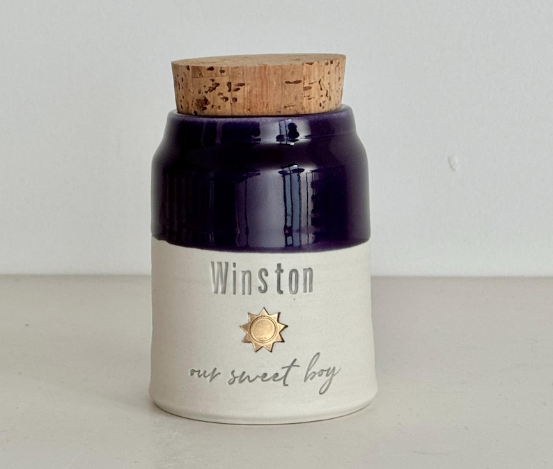 Image shows a collared shape urn with cork lid. It has a dark purple glazed upper section, and white unglazed clay lower portion. There is a gold sun stamp with name  and custom phrase