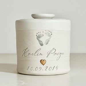 May include: A white ceramic keepsake urn with a lid. The urn features a pair of baby footprints, the name "Kaelin Paige", a gold heart, and the date "10.09.2014". The urn is designed to hold cremated remains.