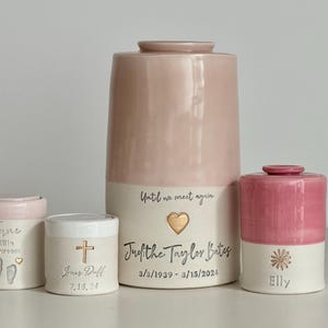 May include: Four ceramic urns in varying sizes, each with a two-tone design of pink and cream. The urns are inscribed with names, dates, and decorative elements like hearts, crosses, and a flower. The largest urn has the text "Until we meet again".