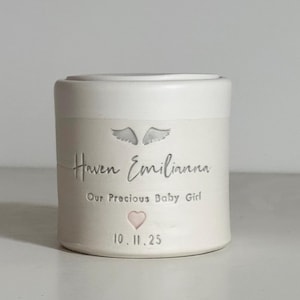 A small white glaze and white porcelain keepsake infant urn. It features a angel wings, the custom name, a phrase, a pink heart, and the date.