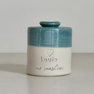 A handmade custom urn with a robins egg blue lid and upper section, and a white lower section made of porcelain clay. The beautiful custom urn features a heart design, the name and a custom phrase