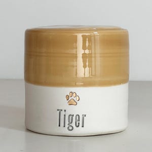 A handmade custom urn with a orange/ wheat color lid and upper section, and a white lower section made of porcelain clay. The beautiful custom urn features a gold paw design, a custom name below