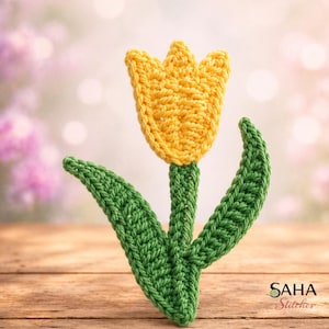 Könnte beinhalten: Eine handgefertigte Häkeltulpe in leuchtendem Gelb und Grün. Die Blume hat ein strukturiertes Stichmuster mit einer gelben Blüte und grünen Stiel und Blättern. Das Bild ist vor einem verschwommenen Hintergrund und einer Holzoberfläche platziert. Das Logo "SAHA Stitches" ist sichtbar.