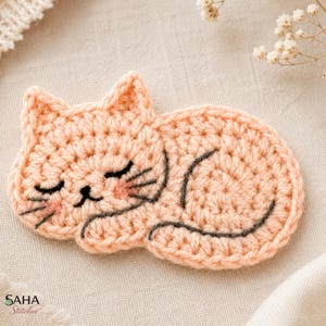 May include: A peach-colored crochet cat applique featuring a sleeping expression with closed eyes, a small black nose, and dark gray embroidered whiskers and tail. The cat has soft pink blush details on its cheeks and the text SAHA Stitches.