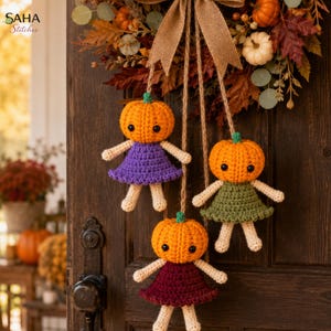 May include: Three crocheted pumpkin-headed dolls hanging from a burlap bow on a wreath. Each doll has a round orange pumpkin head with black eyes, cream-colored arms and legs, and a dress in purple, olive green, or dark red. Text reads SAHA Stitches.