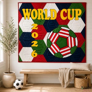 May include: A quilted wall hanging featuring a hexagonal pattern in red, white, blue, and green. The text WORLD CUP 2026 is displayed in large yellow letters. A central soccer ball graphic features red and white stripes on a green background.