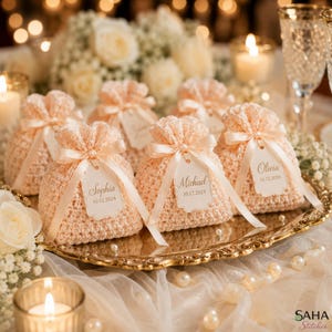 May include: Peach-colored crocheted favor bags with satin ribbon ties and personalized tags. The tags display names and dates, arranged on a gold-toned tray, ideal for weddings or special events.