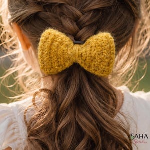 May include: A close-up of a yellow knitted bow hair accessory. The bow is attached to a black hair tie and is positioned on the back of a person's head, with long brown hair in a braid and loose. The bow is made of a textured yarn.