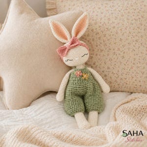 Puede incluir: Una muñeca de conejito de ganchillo hecha a mano con cabeza color crema, orejas rosas y un lazo rosa. La muñeca lleva un mono verde con detalles de flores. Está sobre una cama blanca, con una almohada en forma de estrella y una almohada floral en el fondo.