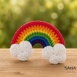 May include: A crocheted rainbow with red, orange, yellow, green, blue, and purple stripes. Two white crocheted clouds are attached at the ends of the rainbow. The rainbow is displayed on a wooden surface.