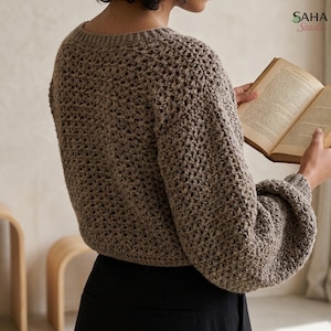 May include: A beige long-sleeved crochet sweater with a textured open-stitch pattern, a round neckline, and slightly puffed sleeves. The garment is worn over black trousers. The text SAHA Stitches is visible in the upper right corner.