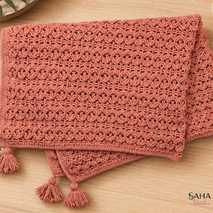 May include: A terracotta-colored crochet blanket featuring a repeating diamond and openwork stitch pattern. The rectangular textile is finished with small matching tassels at the corners and edges, showcasing a detailed, textured yarn construction.
