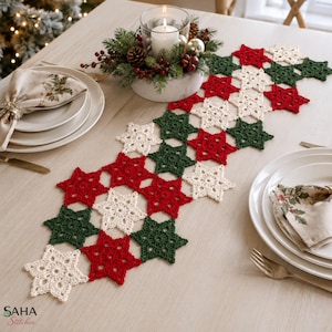 Puede incluir: Un camino de mesa de ganchillo compuesto por motivos en forma de estrella interconectados en hilo rojo, verde oscuro y crema. El camino presenta un patrón repetitivo de estas estrellas, creando una pieza textil festiva para la mesa.