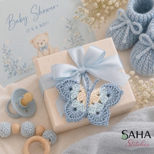 May include: A baby shower gift set with a light blue theme. The set includes a gift box with a blue ribbon, a crocheted butterfly, baby booties, a pacifier, and a wooden teether. A card reads "Baby Shower. It's a Boy!"
