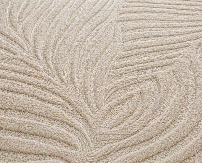 May include: Close-up of a beige rug with a leaf pattern. The design features curved lines that create a textured, three-dimensional effect. The rug's neutral color and detailed pattern suggest a focus on home decor.