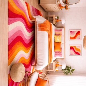 May include: A bedroom scene with a vibrant, wavy rug in shades of red, pink, and orange. The bed has white linens and orange accents, complemented by matching pillows and artwork. A woven chair and a wooden dresser complete the colorful, retro-inspired decor.