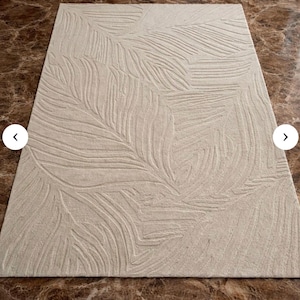 May include: Beige rectangular area rug with a raised leaf pattern. The rug has a soft, neutral color and a textured design, adding a touch of nature-inspired elegance to any room. The rug is made of a durable material.