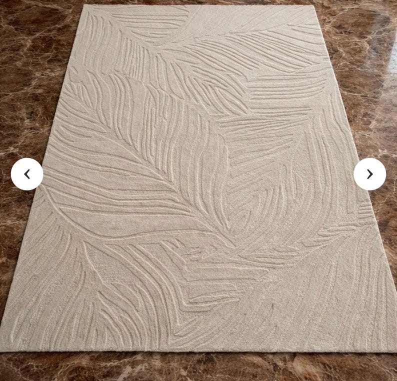 May include: Beige rectangular area rug with a raised leaf pattern. The rug has a soft, neutral color and a textured design, adding a touch of nature-inspired elegance to any room. The rug is made of a durable material.