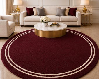 May include: A round, burgundy rug with a cream-colored double border. The rug is placed in a living room setting, with a sofa, coffee table, and lamps visible in the background. The rug's diameter is approximately 8 feet.