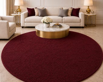 May include: A large, round, burgundy-colored rug dominates the foreground of a living room scene. The rug's texture appears soft and plush. A white sofa with decorative pillows, a marble-topped coffee table, and side tables with lamps complete the elegant setting.