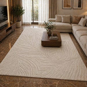 May include: A large, rectangular, off-white rug with a leaf pattern in a living room setting. The rug is placed in front of a sectional sofa and a wooden coffee table. The room has a neutral color palette with natural light.