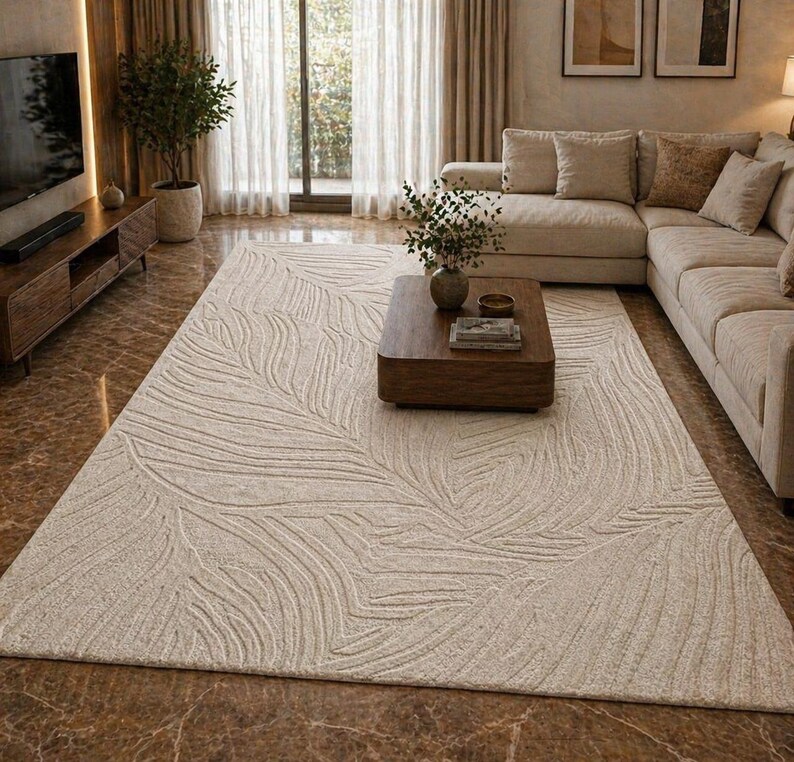 May include: A large, rectangular, off-white rug with a leaf pattern in a living room setting. The rug is placed in front of a sectional sofa and a wooden coffee table. The room has a neutral color palette with natural light.