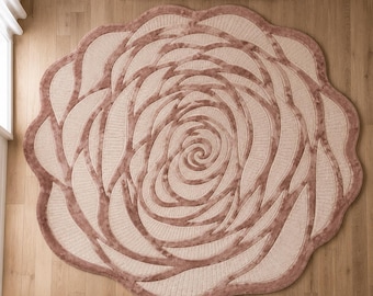 May include: A circular area rug featuring a layered, rose-like floral pattern. The design consists of alternating bands of soft, dusty rose-colored velvet-textured material and lighter cream-colored sections, creating a dimensional, petal-like effect.