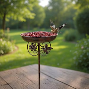 May include: A metal bird feeder with a bowl filled with red seeds. The feeder has a brown finish with floral and hummingbird details. A bird is perched on the edge of the bowl. The feeder is on a tall, slender pole, perfect for a garden.