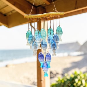 Peut inclure: Un carillon éolien suspendu avec de nombreux poissons en verre bleus et turquoise. Les poissons sont suspendus à une barre en bois. L'arrière-plan montre une plage et l'océan, évoquant un thème côtier.