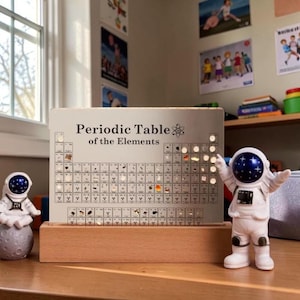 May include: A periodic table of the elements on a light-coloured rectangular board, with the text "Periodic Table of the Elements". The board is set on a wooden base. Two astronaut figurines are on either side of the board.