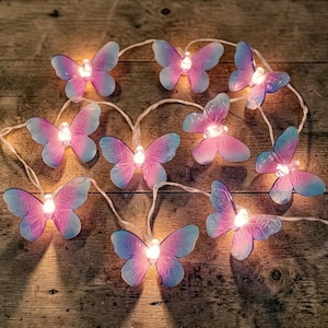 Pode incluir: Uma sequência de luzes decorativas com ornamentos em forma de borboleta. Cada borboleta tem um gradiente de tons rosa e azul, com uma pequena luz no centro. As luzes estão em um fio branco, perfeitas para um toque caprichoso.