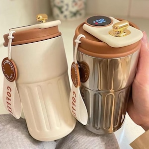 Op de afbeelding: Twee geïsoleerde koffiebekers. Eén is gebroken wit, de andere is roestvrij staal. Beide hebben bruine deksels en gouden accenten. Elk heeft een label met de tekst "COFFEE". De roestvrijstalen beker heeft een digitale temperatuurweergave.