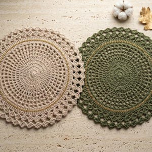 May include: Two round crochet placemats, one in beige and one in olive green, each featuring a central circular pattern, a thin metallic gold thread accent, and a decorative openwork scalloped border edge, displayed side by side.
