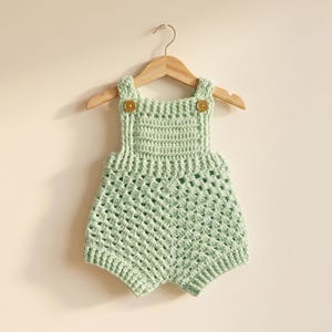 May include: A light green crochet romper with a textured open weave design, hanging on a wooden hanger. The romper features button closures on the straps. The garment is designed for a baby or toddler.