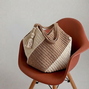Puede incluir: Un bolso de ganchillo con un diseño de bloques de color en secciones diagonales alternas de hilo crema y tostado. El bolso incluye dos asas de ganchillo a juego y una borla decorativa de hilo unida a un lado, mostrando un patrón de punto texturizado.