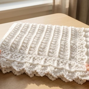 May include: A handmade, white crochet blanket with a textured, ribbed design. The blanket features a scalloped edge and is displayed on a wooden table. The soft, off-white color and intricate stitching create a cozy, handcrafted feel.