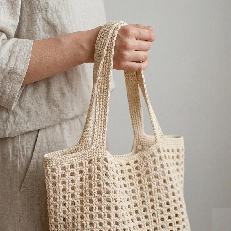 Pu&ograve; includere: Una borsa tote all'uncinetto color crema caratterizzata da un motivo a griglia traforata su tutto il corpo. La borsa &egrave; dotata di due robusti manici a maglia fitta che si estendono dal bordo superiore, offrendo una consistenza uniforme.