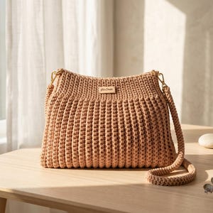May include: A beige crochet shoulder bag with a rectangular shape and a long, braided strap. The bag features a small, rectangular label with text. The bag is sitting on a light-colored wooden table.