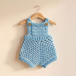 May include: A light blue crochet romper for a baby, hanging on a wooden hanger. The romper has a textured stitch pattern and is secured with two brown buttons on the straps. The garment is displayed against a neutral background.