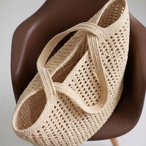 Pu&ograve; includere: Una borsa tote all'uncinetto color beige caratterizzata da un motivo a rete a maglia aperta su tutto il corpo. La borsa presenta due manici integrati e robusti che si incrociano sopra l'apertura superiore, creando una silhouette strutturata.