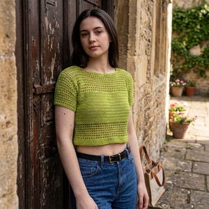 May include: A lime green short-sleeved crochet crop top featuring a round neckline and a combination of solid and open-work stitch patterns. The garment is paired with blue denim jeans secured by a black leather belt with a silver-toned metal buckle.