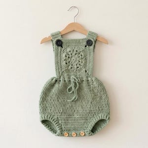 May include: Sage green crochet baby romper with a round, openwork design on the bib. The romper has adjustable straps with black buttons, a drawstring waist, and wooden buttons at the bottom. It hangs on a wooden hanger.