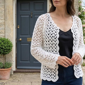May include: A white, open-weave crochet cardigan with a scalloped edge. The long-sleeved garment is worn over a black top and blue jeans. The cardigan's intricate pattern is visible, creating a delicate, airy look. The background includes a blue door and greenery.