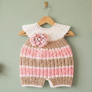 May include: A crochet romper featuring a white yoke with a ruffled edge, followed by horizontal stripes in alternating tan and light pink. A decorative bow with a patterned print and a central pearl accent is attached to the upper chest area.
