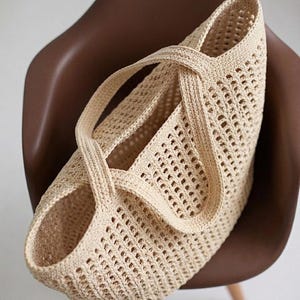 Pu&ograve; includere: Una borsa tote all'uncinetto color beige caratterizzata da un motivo a rete a maglia aperta su tutto il corpo. La borsa presenta due manici integrati e robusti che si incrociano sopra l'apertura superiore, creando una silhouette strutturata.