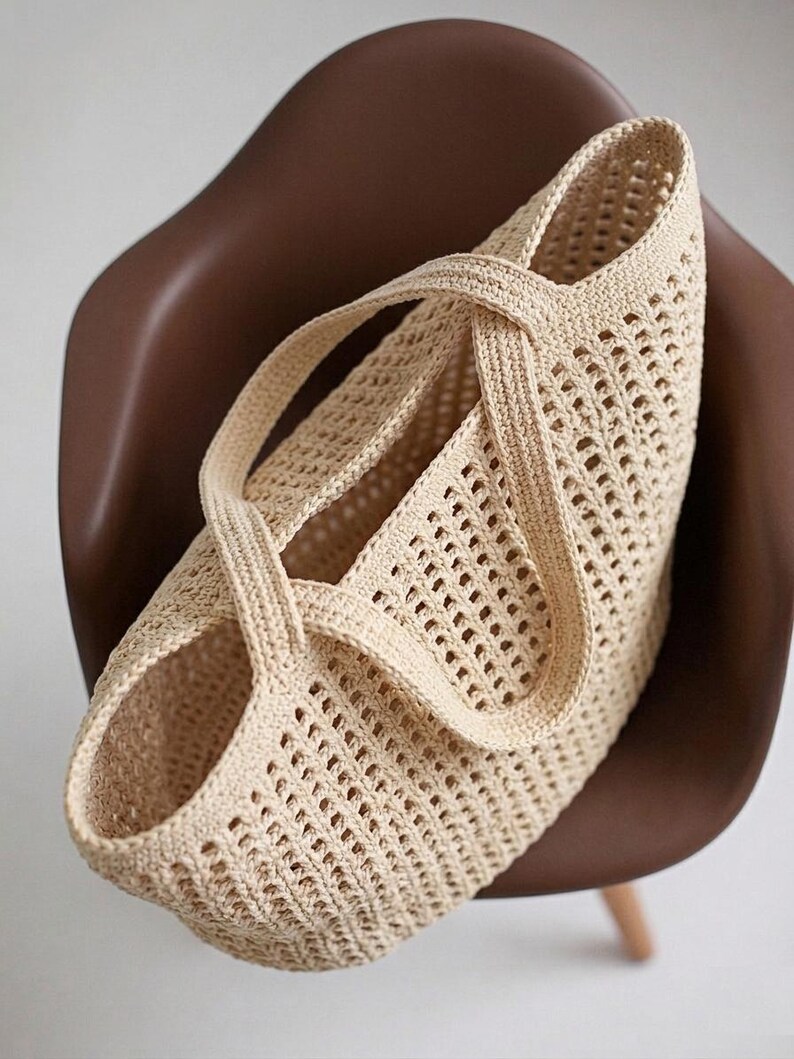 Pu&ograve; includere: Una borsa tote all'uncinetto color beige caratterizzata da un motivo a rete a maglia aperta su tutto il corpo. La borsa presenta due manici integrati e robusti che si incrociano sopra l'apertura superiore, creando una silhouette strutturata.